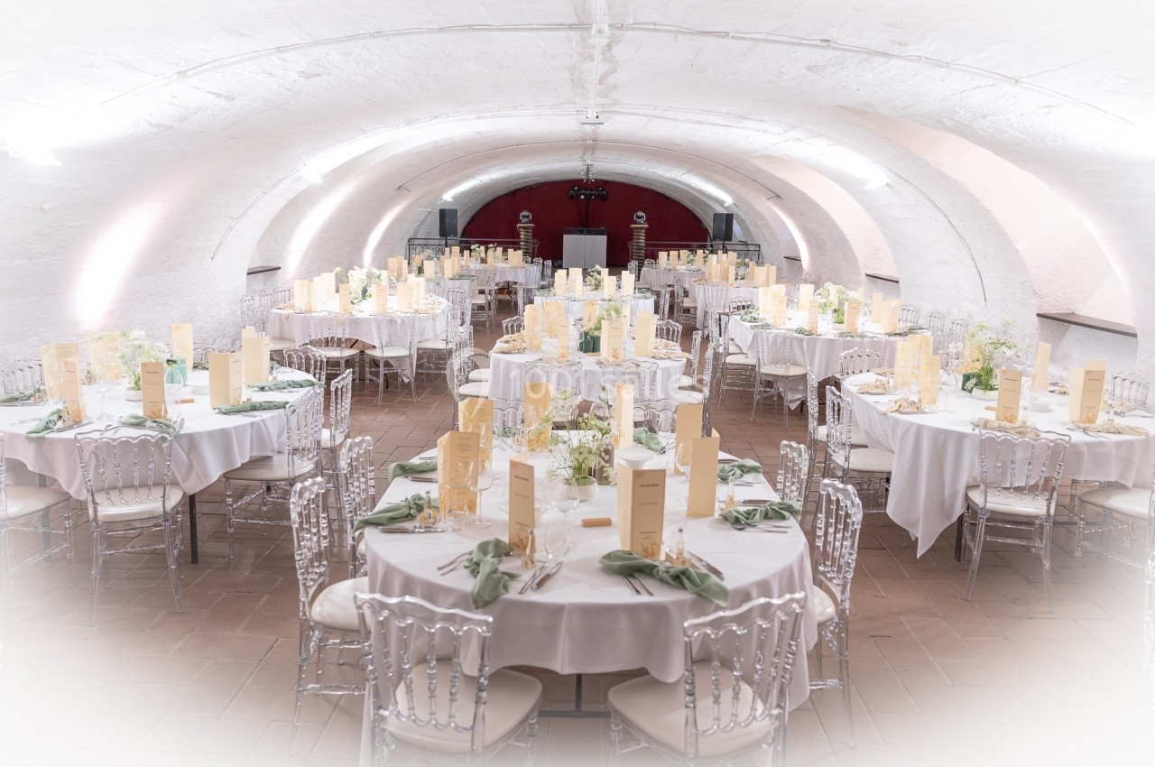 Salle voûtée décorée pour un banquet, avec tables rondes dressées, nappes blanches et chaises transparentes.