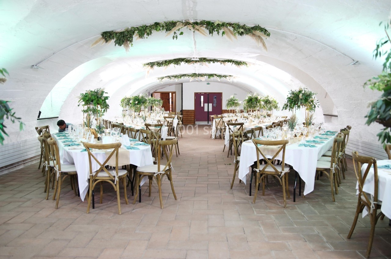 Salle voûtée décorée pour un événement, avec tables dressées, chaises en bois et ornements végétaux suspendus.