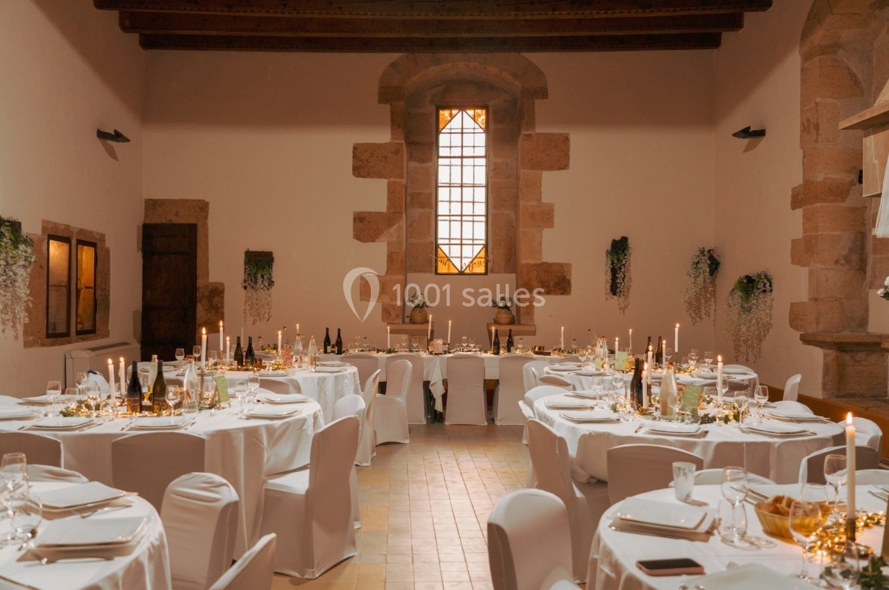 Salle de réception décorée avec des tables rondes, nappes blanches, chandeliers et lumière naturelle provenant d'une grande…