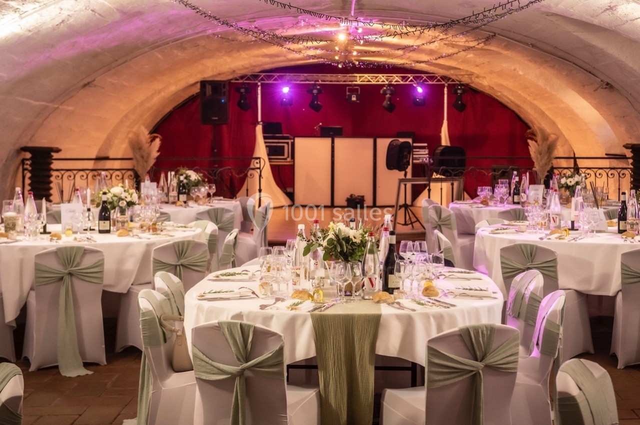Salle voûtée décorée pour un événement, avec tables rondes dressées, nappes blanches et chaises ornées de rubans verts.