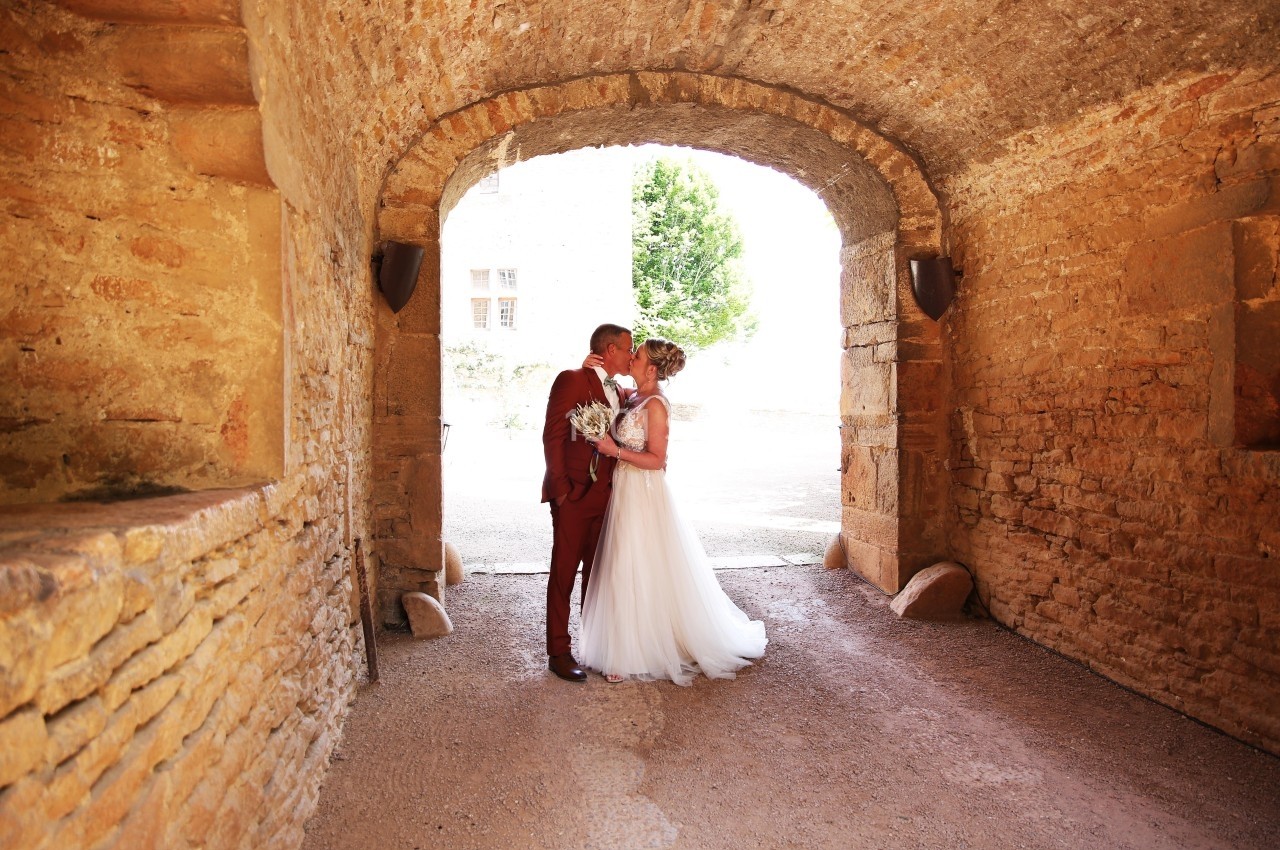 Un couple de mariés s'embrasse sous une arche en pierre, baignée de lumière naturelle.