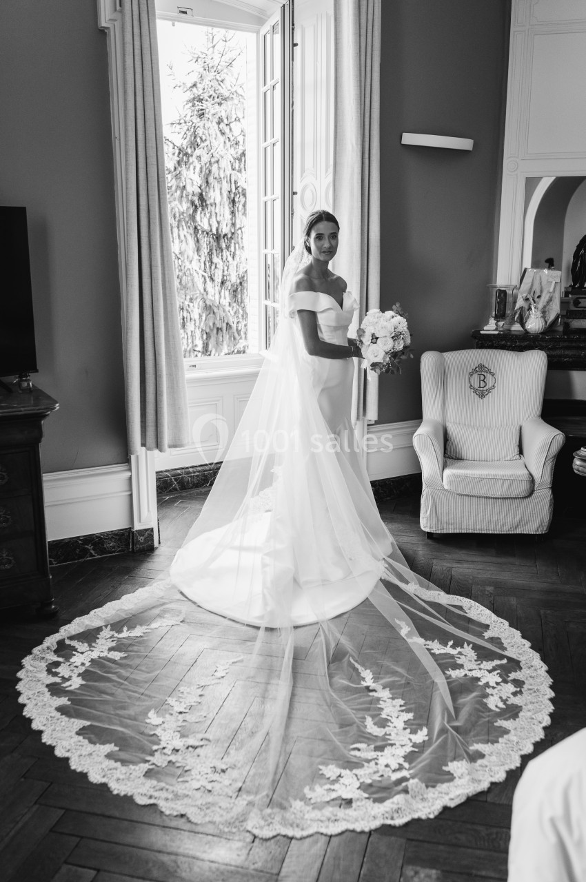 Une mariée en robe blanche avec un long voile en dentelle se tient dans une pièce élégante près d'une fenêtre.
