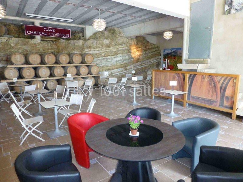 Salle de dégustation avec tables, fauteuils et tonneaux en bois alignés contre un mur de pierre.