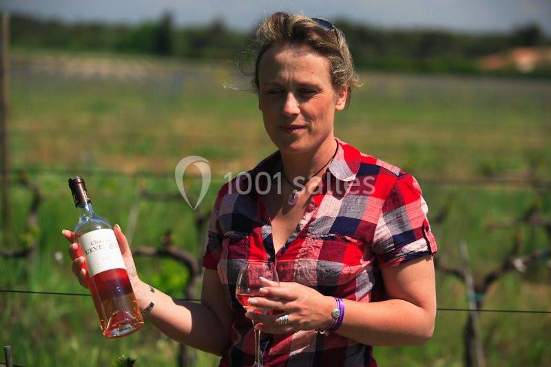 Femme tenant une bouteille de vin rosé et un verre, debout dans un vignoble par une journée ensoleillée.