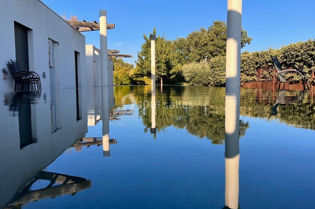 Reflet d'une maison moderne et de végétation sur une surface d'eau calme sous un ciel bleu.