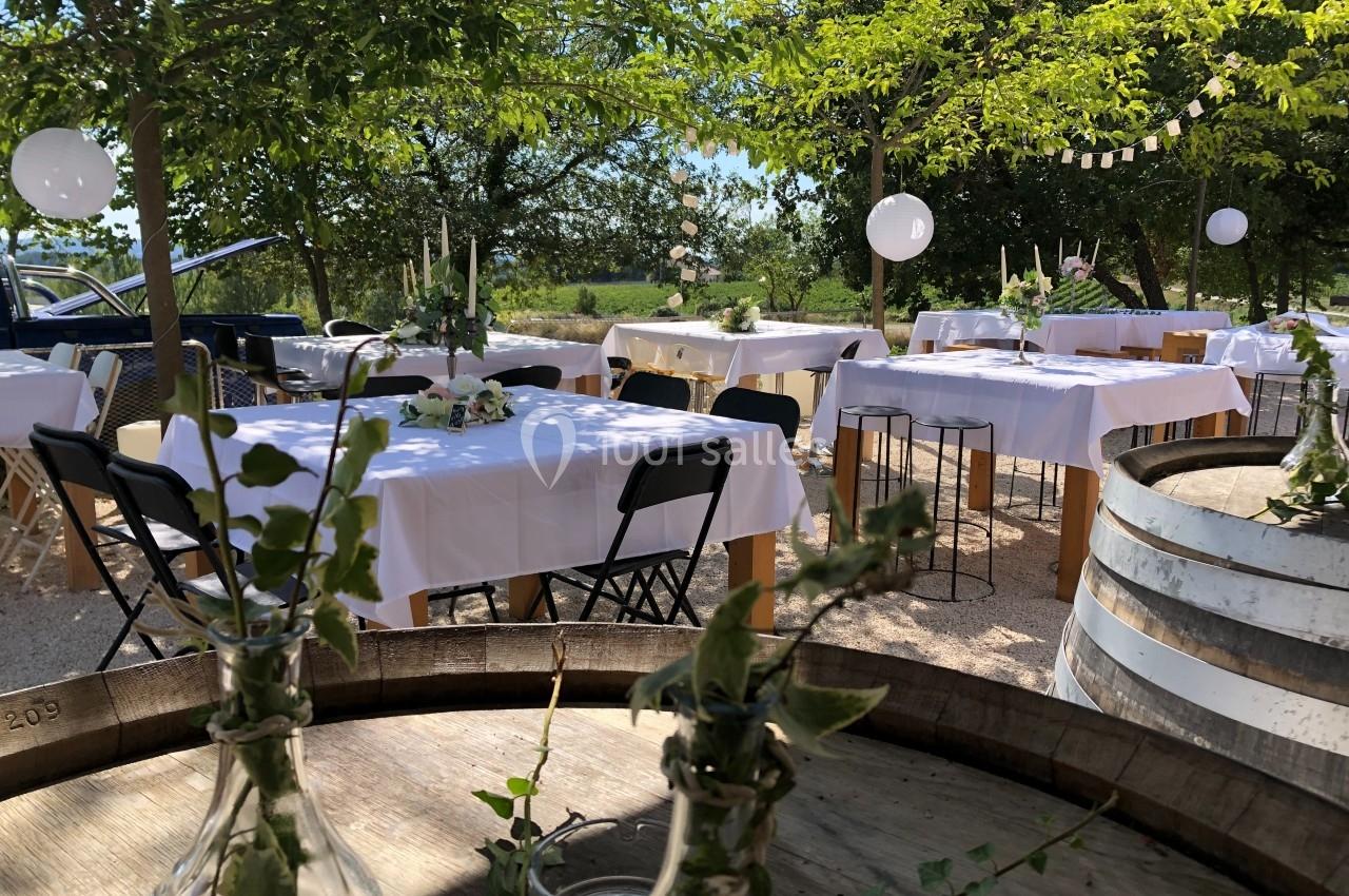 Tables décorées avec nappes blanches et bouquets, disposées en extérieur sous des arbres pour un événement festif.