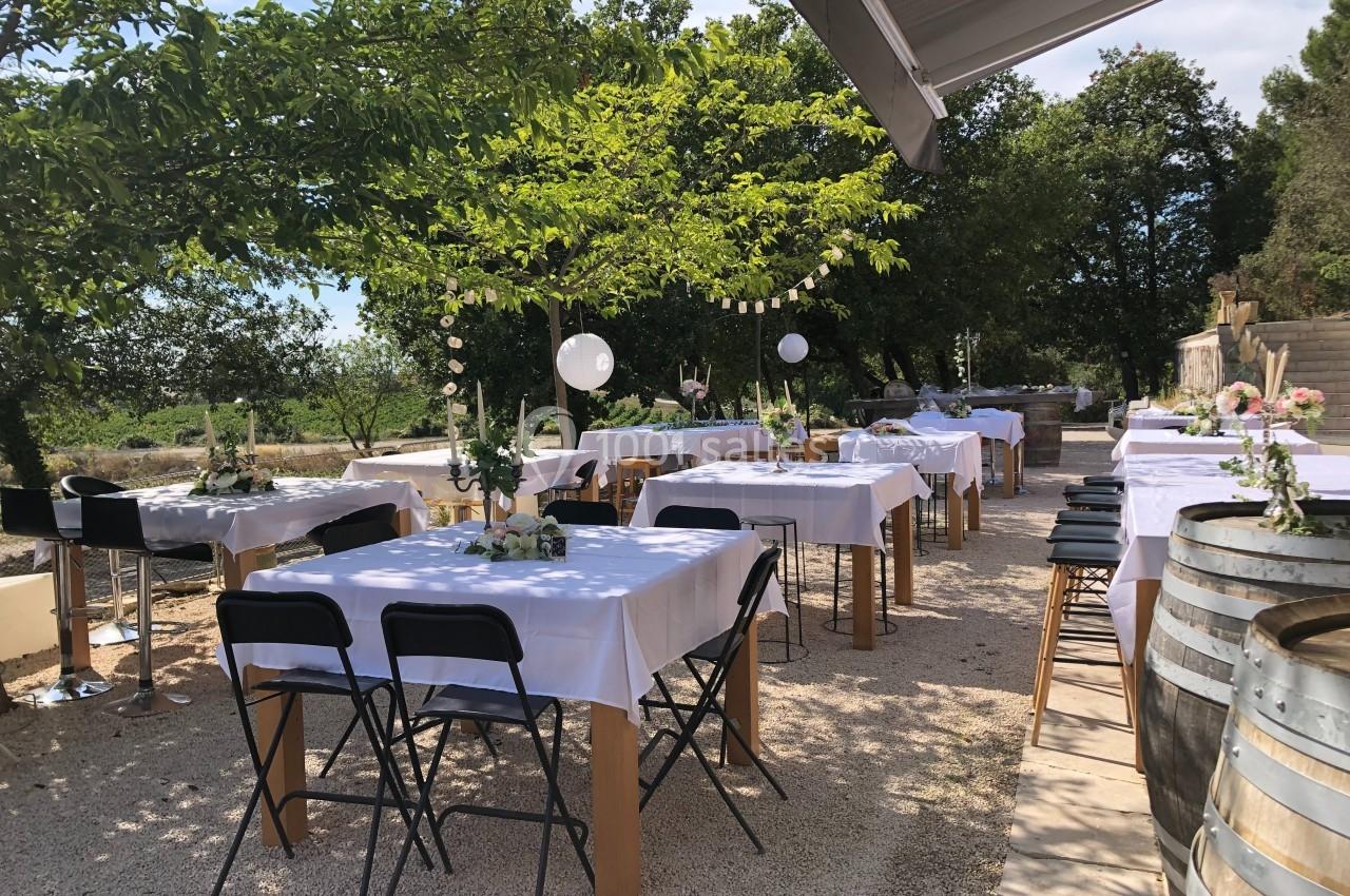 Tables dressées avec nappes blanches sous des arbres, décorées pour un événement en extérieur par temps ensoleillé.