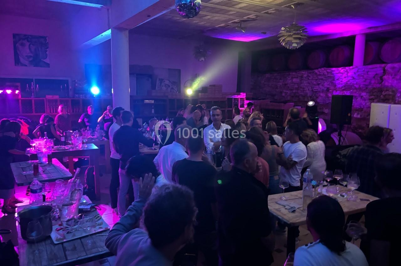 Groupe de personnes rassemblées dans une salle éclairée par des lumières colorées, avec tables et tonneaux en arrière-plan.