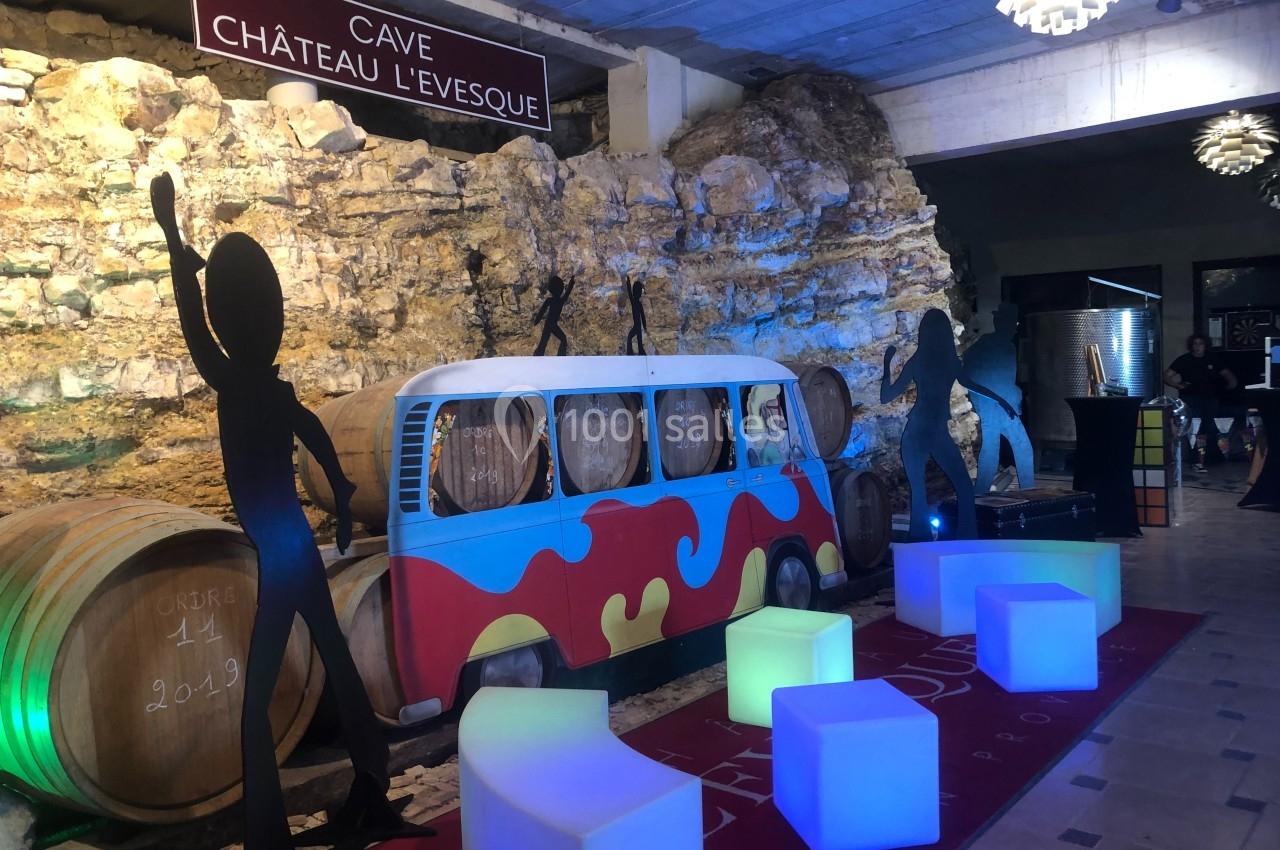 Décor de cave avec tonneaux, combi vintage coloré, silhouettes noires et cubes lumineux dans une ambiance festive.