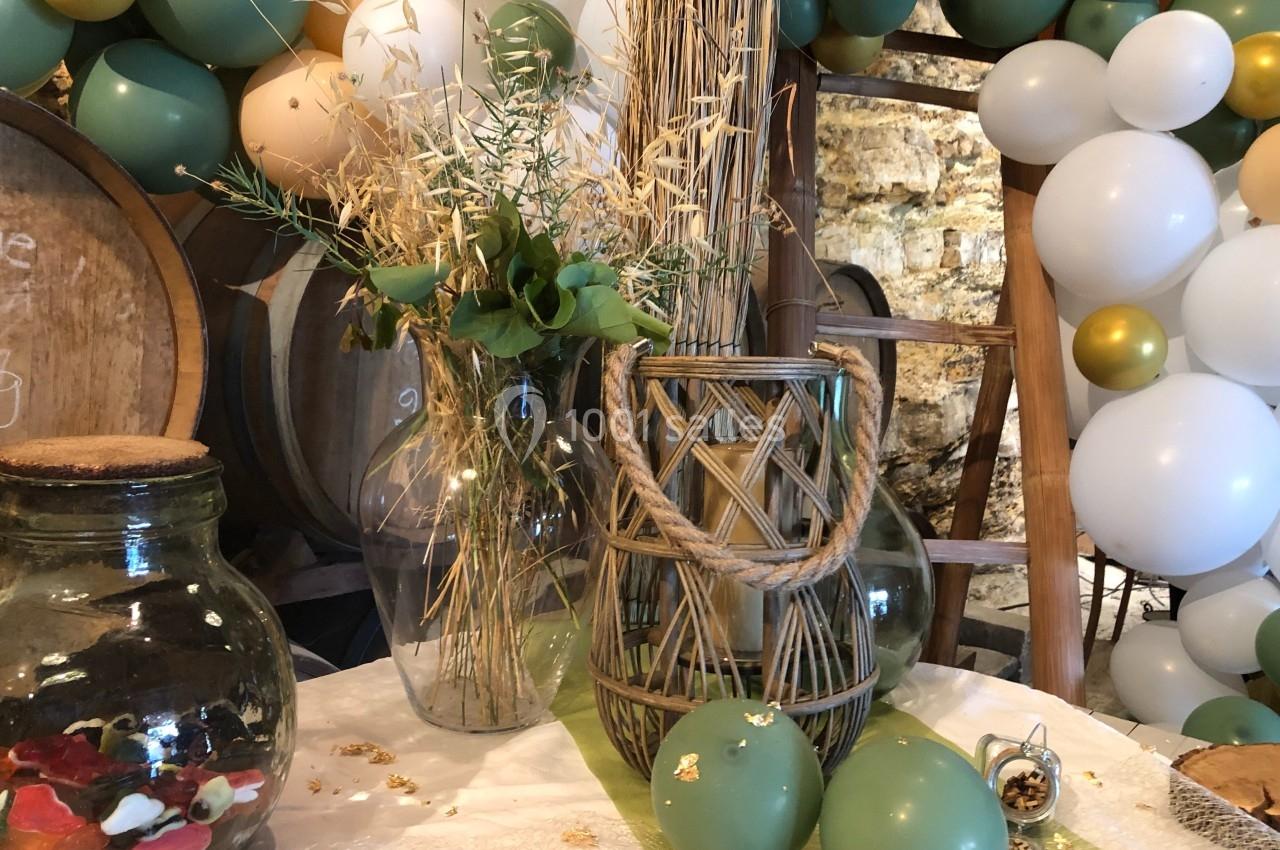 Décoration de table avec vases en verre, panier en osier, ballons verts et blancs, et éléments naturels.