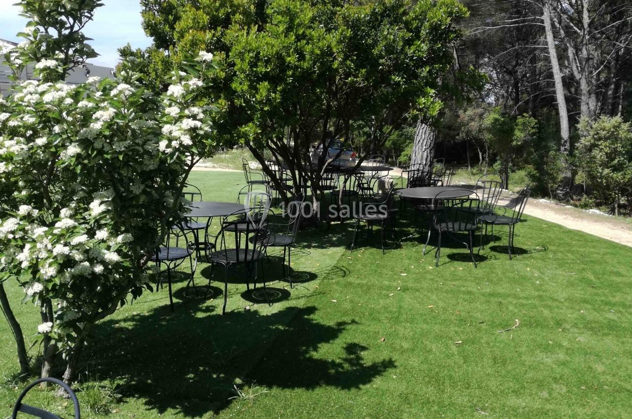 Tables et chaises en métal noir disposées sur une pelouse ombragée par des arbres et arbustes en fleurs.