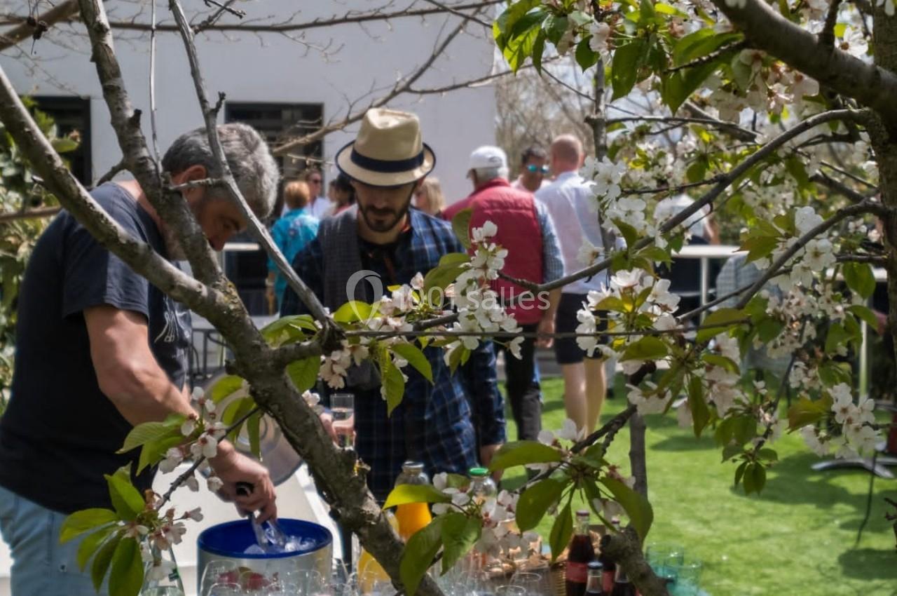Des personnes se servent à une table garnie de boissons et snacks dans un jardin ensoleillé, entouré d'arbres en fleurs.