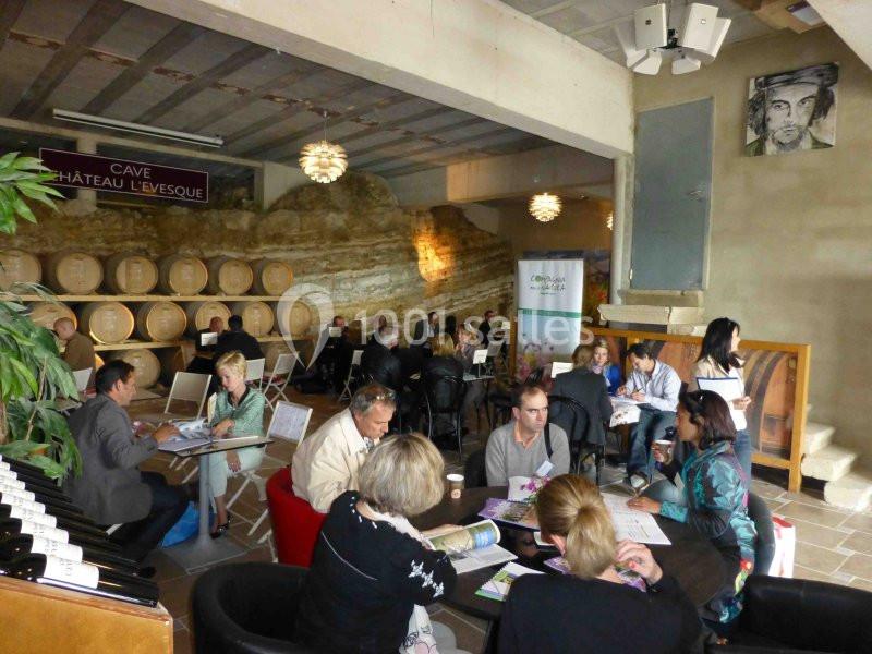 Des personnes discutent autour de tables dans une cave avec des tonneaux en arrière-plan et un mur de pierre visible.