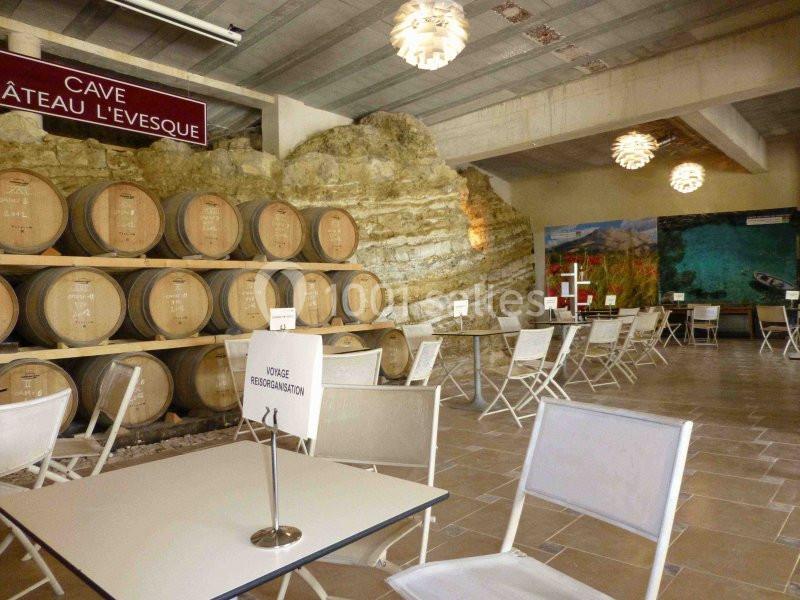 Salle de dégustation avec tables, chaises pliantes et tonneaux de vin alignés contre un mur de pierre apparente.