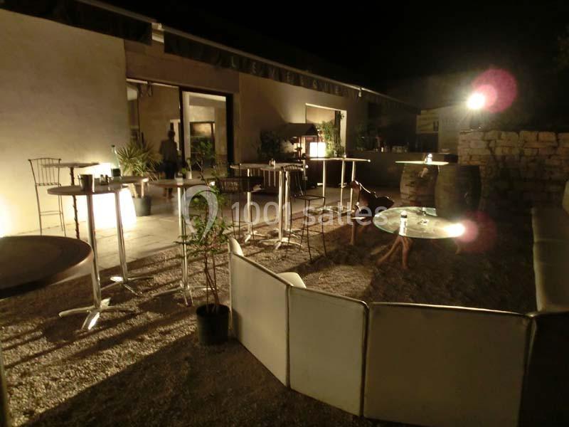 Terrasse extérieure éclairée de nuit avec tables hautes, chaises et canapés disposés sur un sol en gravier.