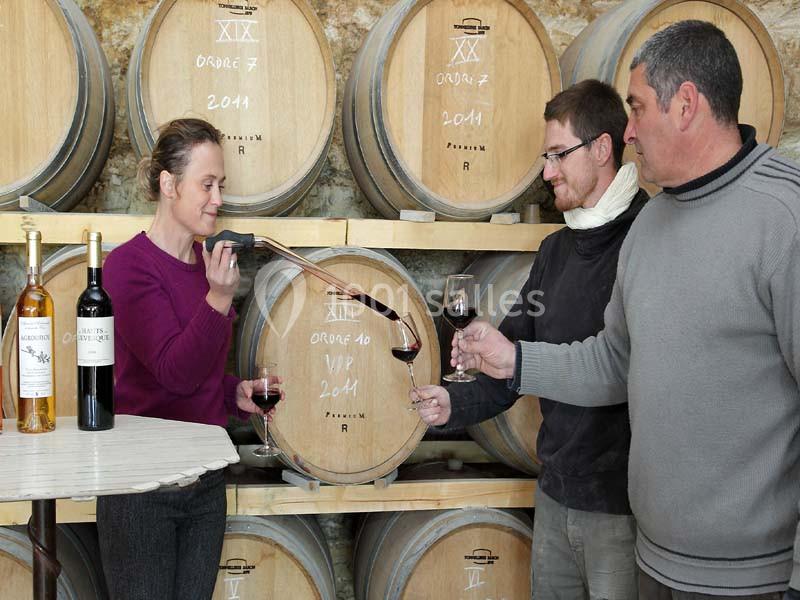 Une femme sert du vin rouge à deux hommes dans une cave avec des tonneaux en arrière-plan.