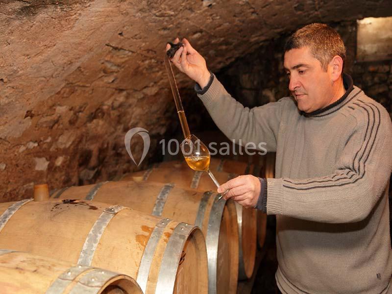 Un homme prélève du vin avec une pipette dans un verre au-dessus de fûts en bois dans une cave voûtée.