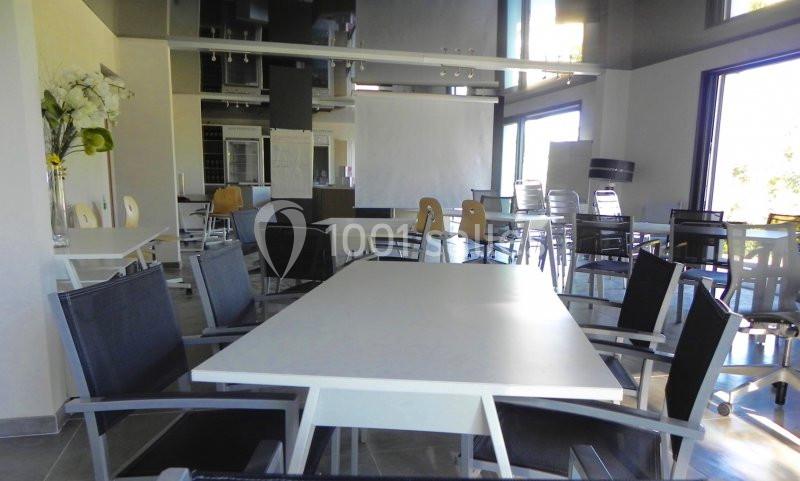 Salle lumineuse avec tables et chaises modernes, grandes fenêtres et écran de projection en arrière-plan.