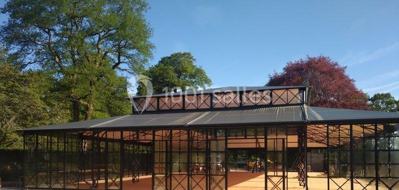 Structure en métal et verre avec toit incliné, entourée d'arbres, sous un ciel bleu dégagé.