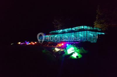 Bâtiment en verre entouré de végétation, avec des arbres et des rochers sous un ciel bleu dégagé.