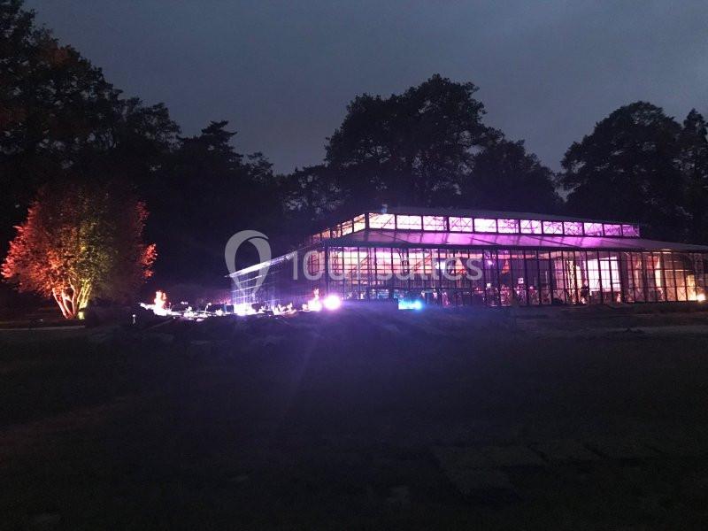 Serre illuminée par des lumières colorées dans un jardin sombre, entourée d'arbres en arrière-plan.