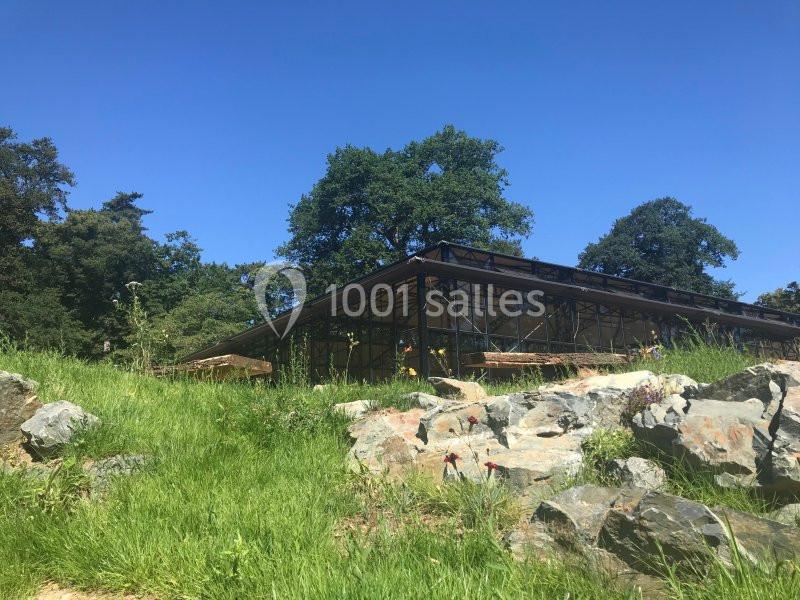 Bâtiment en verre entouré de végétation, avec des arbres et des rochers sous un ciel bleu dégagé.