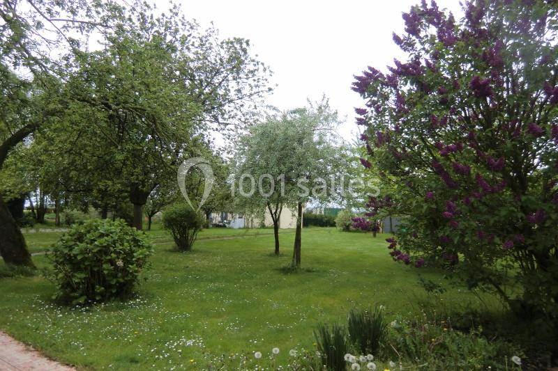 Jardin verdoyant avec pelouse, arbres feuillus et lilas en fleurs, sous un ciel légèrement couvert.