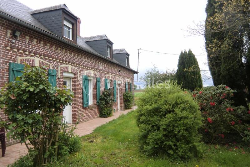 Maison en briques rouges avec volets verts, entourée d'un jardin arboré et fleuri sous un ciel nuageux.