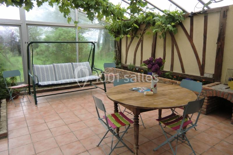 Véranda lumineuse avec table en bois, chaises colorées, canapé suspendu et plantes grimpantes au plafond.