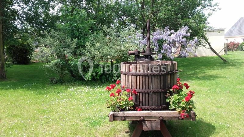 Ancien pressoir en bois décoré de fleurs rouges, placé sur une pelouse verdoyante avec des arbres et arbustes en arrière…