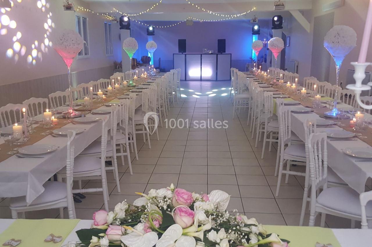 Salle de réception décorée pour un mariage, avec tables alignées, centre de table floral et éclairage festif.
