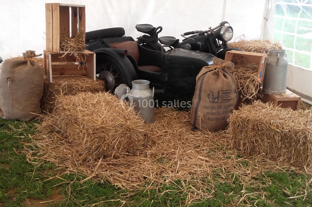 Moto avec side-car entourée de bottes de paille, sacs en toile de jute et bidons en métal, dans une tente sur herbe.