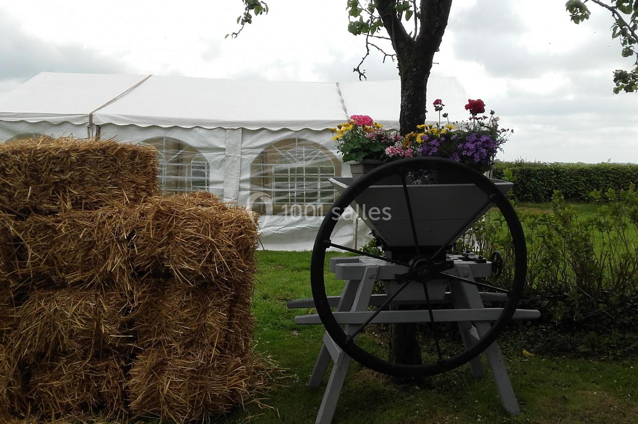 Bottes de paille empilées près d'une charrette fleurie sous un arbre, avec une tente blanche en arrière-plan.