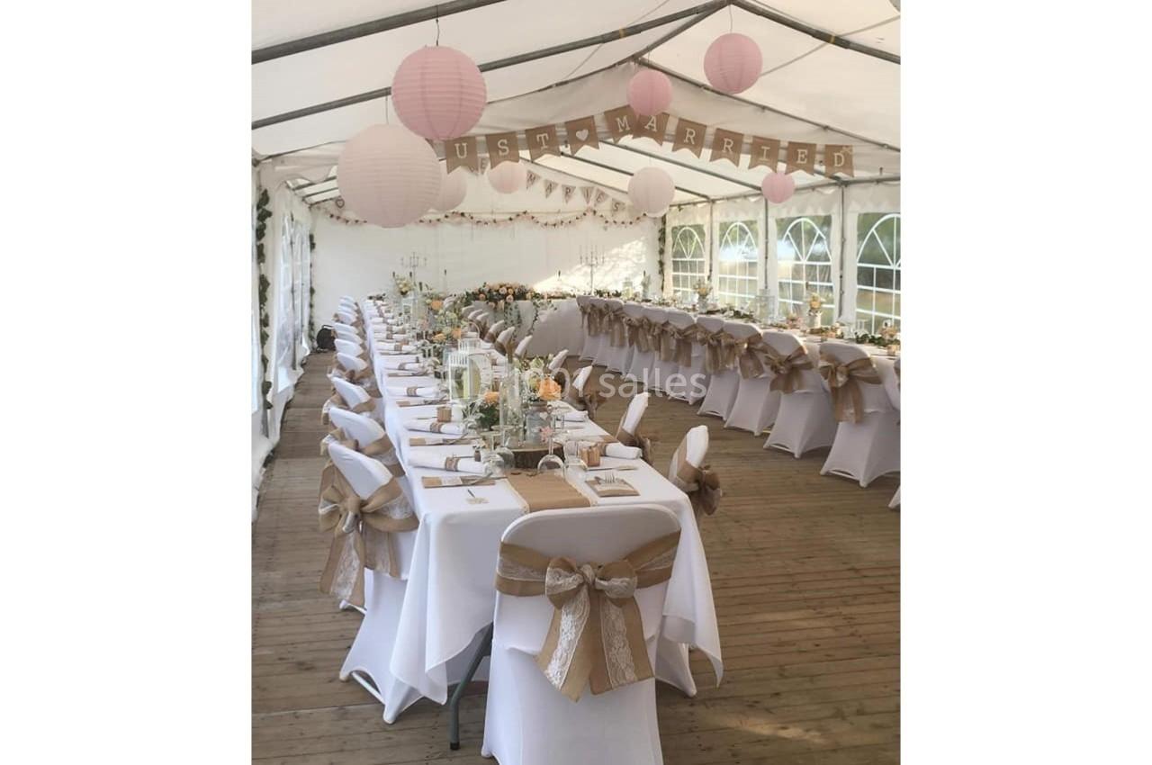 Salle de réception décorée pour un mariage, avec tables alignées, nappes blanches et chaises ornées de rubans beige.