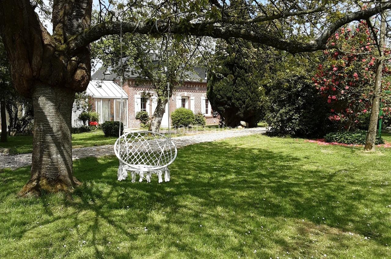 Jardin verdoyant avec une chaise blanche sous un arbre, devant une maison en pierre et des arbustes fleuris.