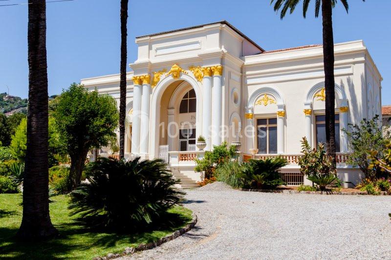 Bâtiment blanc à l'architecture classique avec des détails dorés, entouré de palmiers et d'un jardin verdoyant.