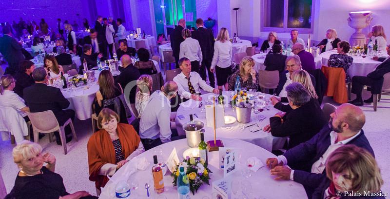 Salle de réception avec des invités assis à des tables rondes, décorée de fleurs et éclairée par une lumière tamisée.