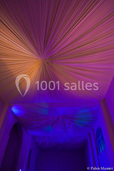 Plafond drapé de tissu beige éclairé par des lumières violettes et bleues dans une salle aux grandes fenêtres.