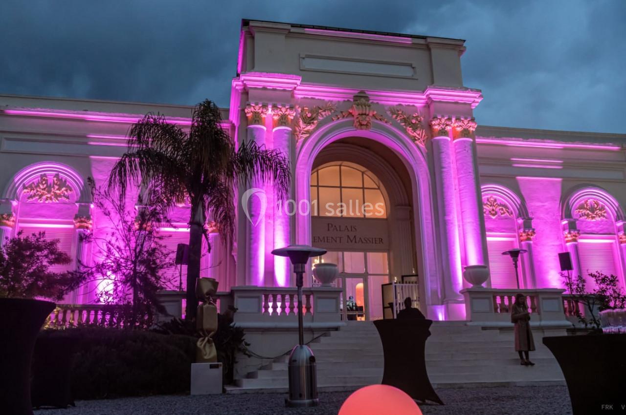 Façade d'un bâtiment éclairé en rose avec des colonnes et une grande arche, vue de nuit avec un ciel nuageux.