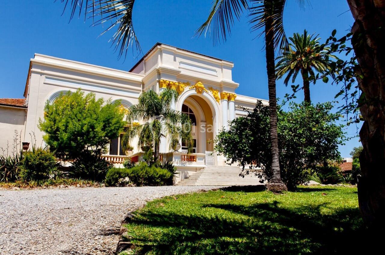 Façade d'un bâtiment blanc avec des arches dorées, entouré de palmiers et d'un jardin sous un ciel bleu.