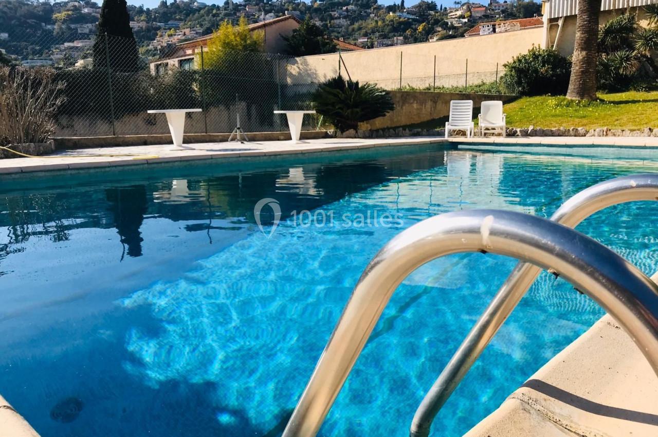 Piscine extérieure avec eau bleue, entourée de verdure et de chaises longues sous un ciel ensoleillé.