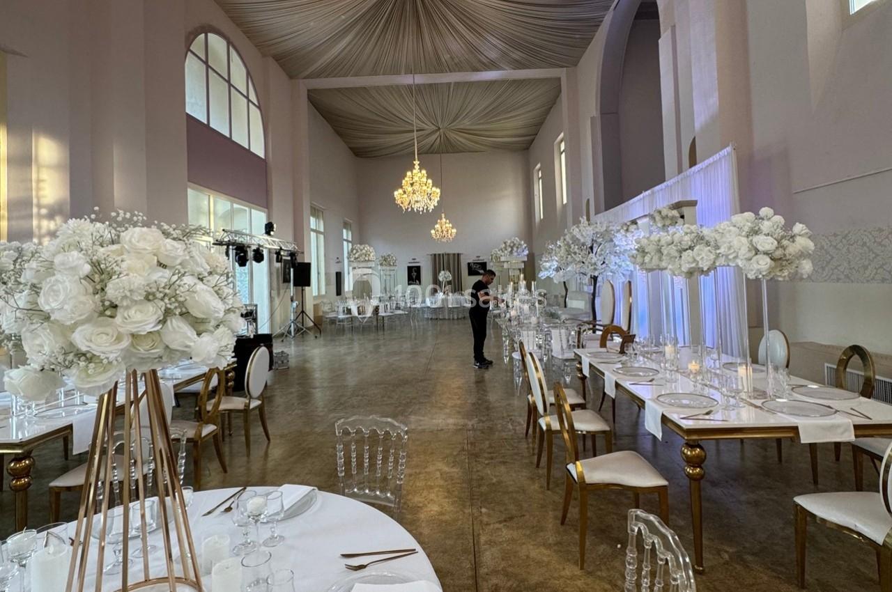 Salle de réception élégante avec tables dressées, chaises transparentes et décorations florales blanches sous un plafond…