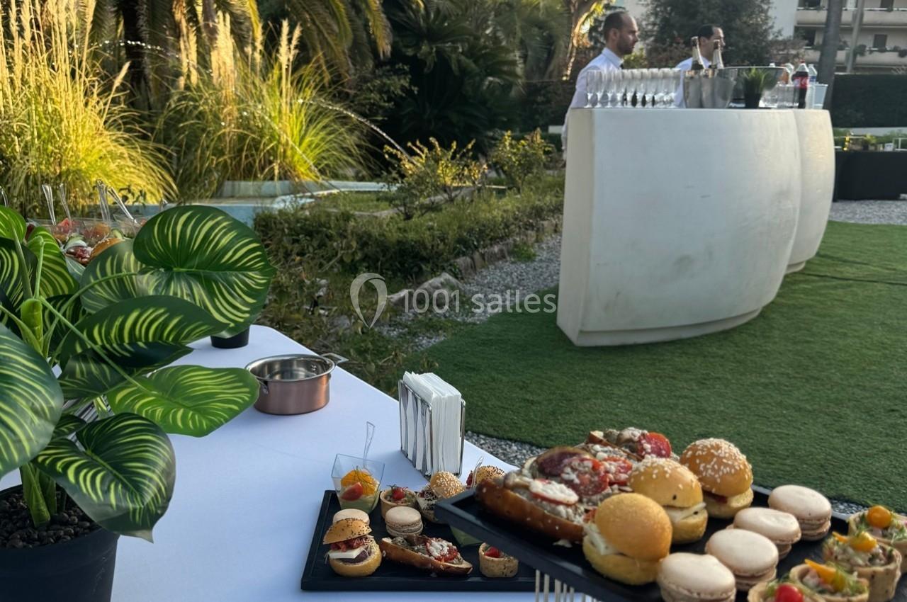 Buffet en plein air avec amuse-bouches variés sur une table, un serveur derrière un bar moderne, et un jardin en arrière…