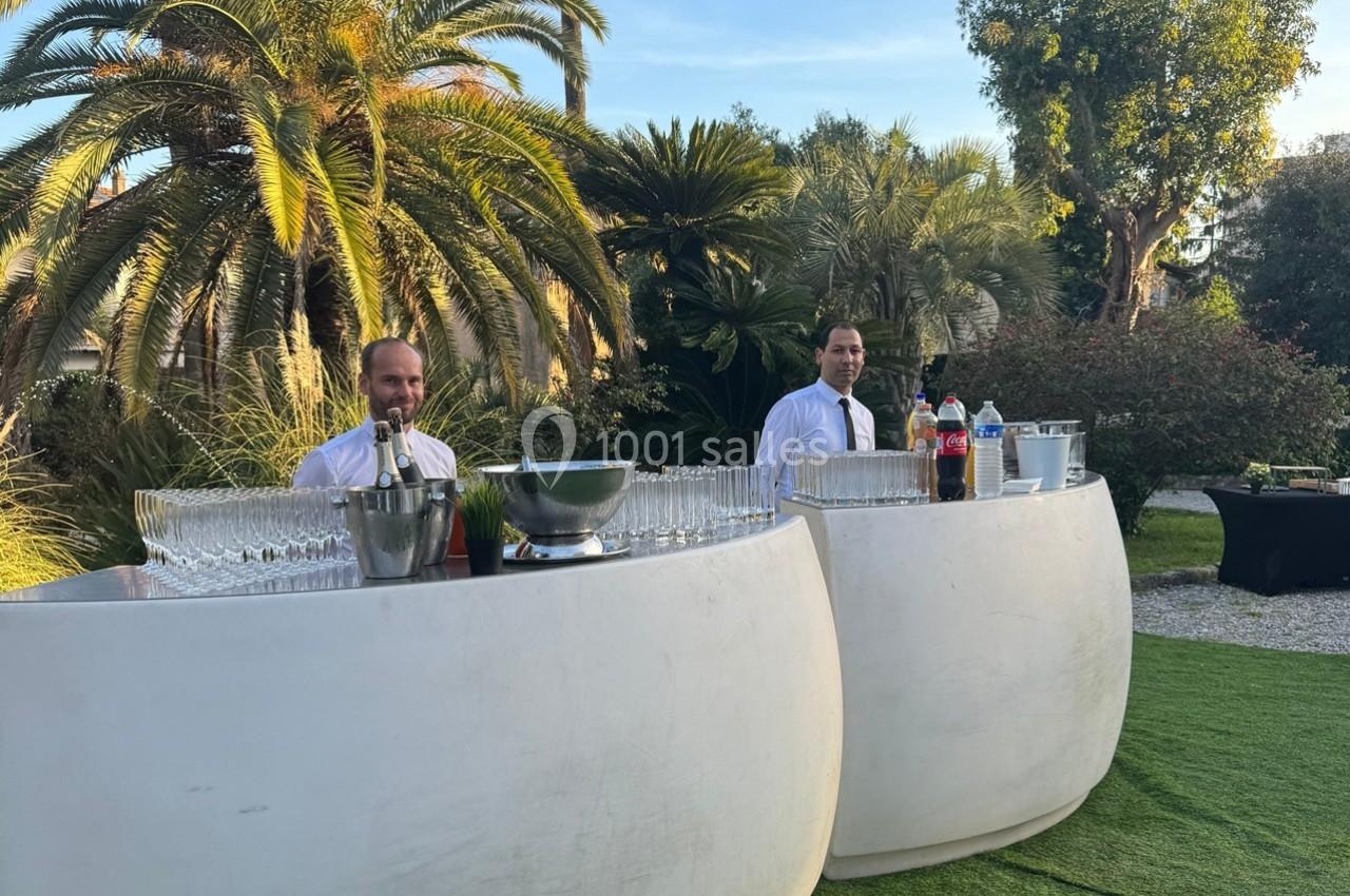 Deux serveurs derrière un bar extérieur avec verres et boissons, entourés de végétation tropicale.