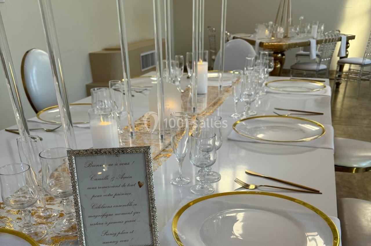 Table élégamment dressée avec des assiettes bordées d'or, des verres, des bougies et un cadre décoratif.