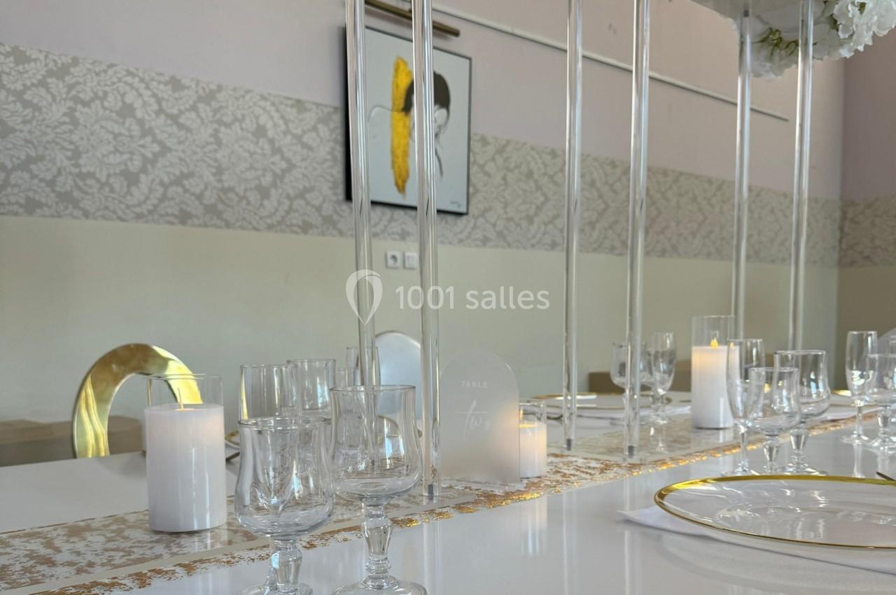 Table élégamment dressée avec verres, assiettes dorées, bougies et décoration florale dans une salle lumineuse.