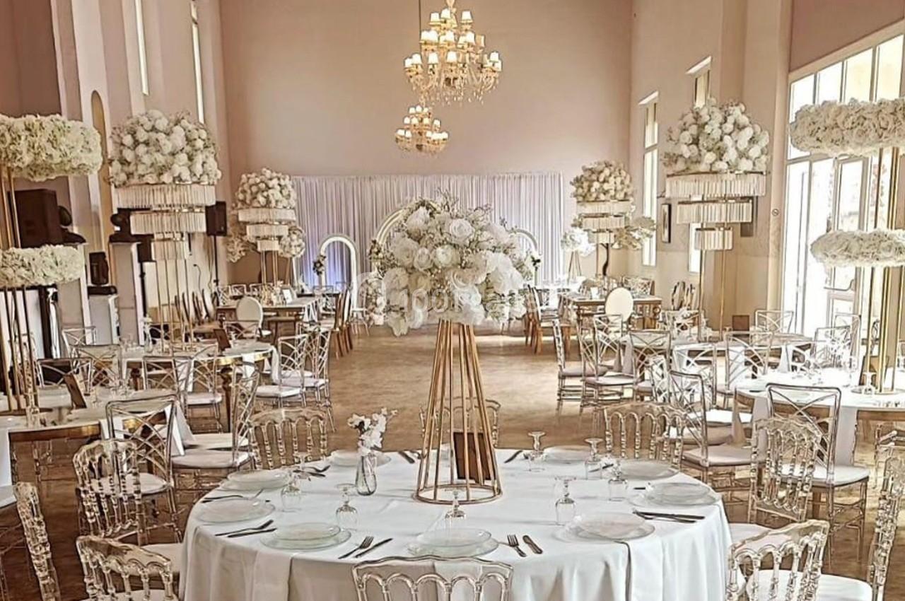 Salle de réception élégante décorée de fleurs blanches, avec tables dressées et chaises transparentes.