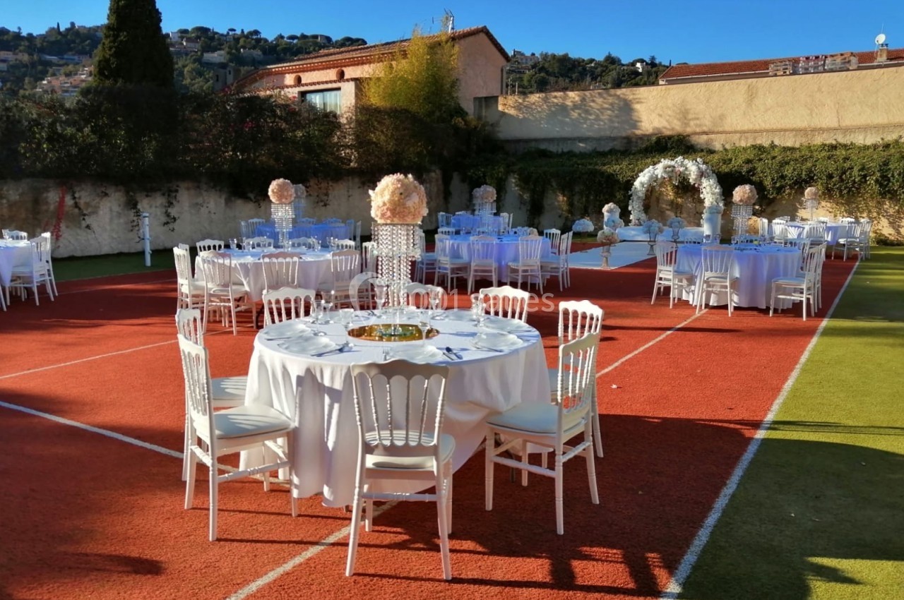 Tables rondes décorées pour un événement en extérieur, disposées sur un terrain de sport avec des arches fleuries.