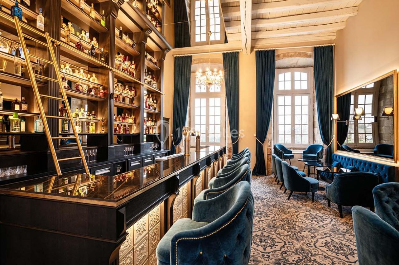 Bar élégant avec étagères remplies de bouteilles, comptoir en bois sombre, fauteuils bleus et grandes fenêtres lumineuses.