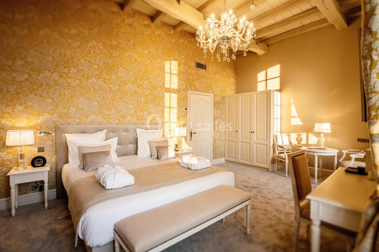 Chambre élégante avec lit double, mobilier clair, lustre, papier peint doré et lumière naturelle entrant par les fenêtres.